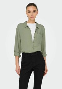 Lichtgroene overhemddas met opgerolde mouwen, zakdetails, gedragen over een witte t-shirt, gecombineerd met zwart hoge taille broeken.