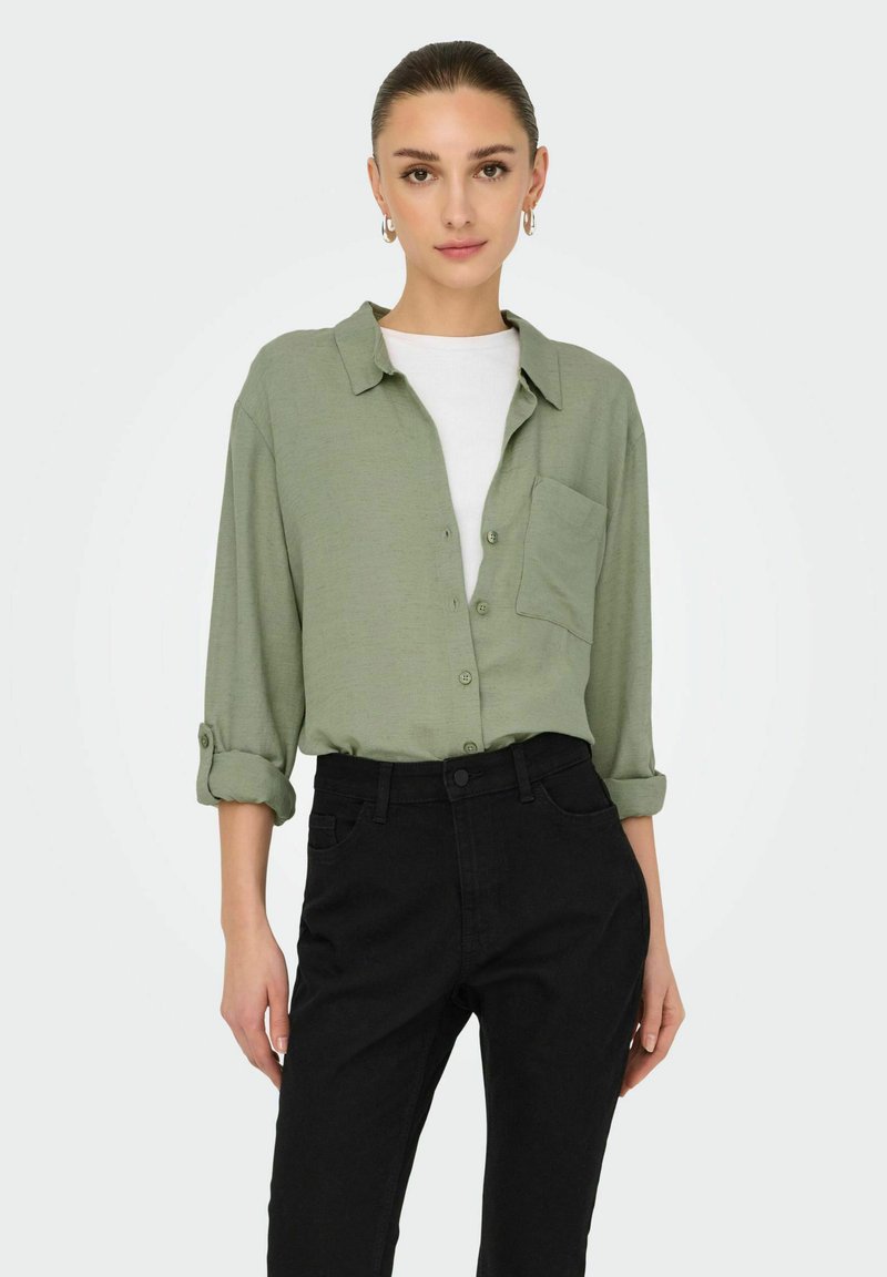 Lichtgroene overhemddas met opgerolde mouwen, zakdetails, gedragen over een witte t-shirt, gecombineerd met zwart hoge taille broeken.