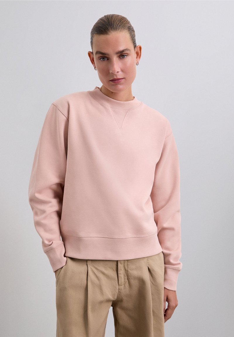 Kvinna med bakdraget hår som bär en ljusrosa sweatshirt och beige byxor, stående mot en enfärgad ljusgrå bakgrund.