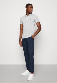 Polo grigia con logo verde, abbinata a pantaloni blu e sneakers bianche. La maglietta presenta un colletto classico e maniche corte.
