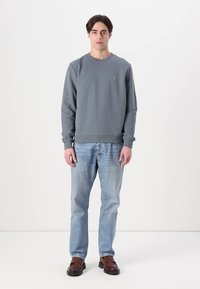 Szürke pulóver kerek nyakkal, bordás mandzsettával és derékpánttal, világoskék farmernadrággal és barna slip-on cipőkkel párosítva. A modellt fehér háttér előtt áll.