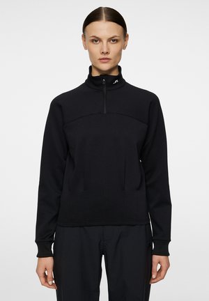 Pull-over zippé noir en tissu doux, avec un col haut, des détails de couture à l'avant et des poignets côtelés. Design minimaliste sans logos visibles.