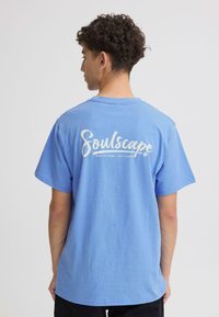 T-shirt en coton bleu avec un grand texte blanc "Soulscape" imprimé dans le dos. Manches courtes et coupe décontractée avec un col rond.