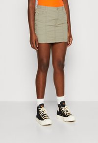 Khaki mini sukně s bočními kapsami, vyrobená z texturované látky. Spárována s černými vysokými teniskami a bílými ponožkami.