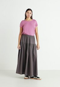 Rosa t-skjorte med korte ermer kombinert med en mørkegrå, lagdelt maxiskjørt. Modellen har på seg svarte sneakers med beige såler. Enkle og uformelle design.