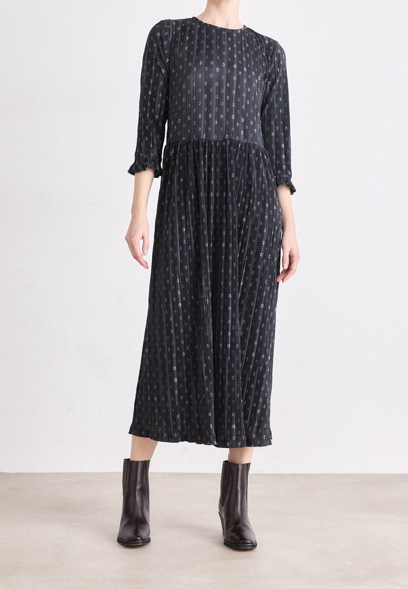 Femme portant une robe midi noire avec de petits motifs blancs et des manches trois-quarts, associée à des bottines foncées.