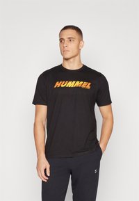 Juoda sportinė marškinė su "HUMMEL" logotipu oranžinės ir geltonos gradientinės spalvos per krūtinę. Apvalia kaklo iškirptė, trumpomis rankovėmis.