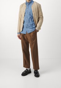 Beżowa pikowana kurtka, niebieska koszula zapinana na guziki, brązowe spodnie typu wide-leg, czarne loafersy z srebrnymi akcentami, noszone z szarymi skarpetkami.