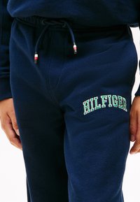 Κοντινό πλάνο χεριών κοντά σε σκούρες μπλε φόρμες με κορδόνι και κεντημένο το "HILFIGER" με πράσινα και άσπρα γράμματα στη δεξιά μηρός.