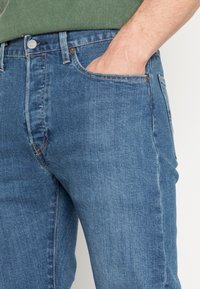 Personne portant un jean en denim bleu avec la main droite partiellement dans la poche avant, chemise verte visible rentrée.