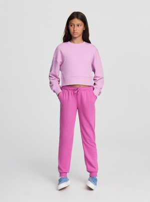 Młoda osoba stojąca z rękami w kieszeniach, ubrana w jasnofioletową krótka bluzę, różowe spodnie dresowe i niebieskie trampki na jednolitym szarym tle.