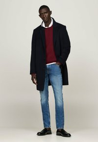 Cappotto navy, maglione bordeaux, camicia bianca, jeans azzurri chiari e scarpe nere. Presenta un design sartoriale e una texture liscia.