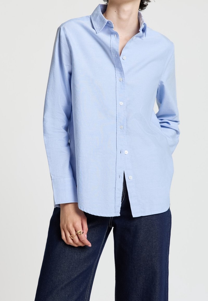 Personne portant une chemise bleu clair à manches longues boutonnée et un jean bleu foncé, une main reposant sur la hanche.
