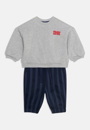 Sweat-shirt gris court à manches longues avec texte rouge sur la poitrine, associé à un pantalon à rayures verticales bleu marine et noir avec cordon de serrage.