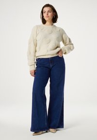 Witte trui met gestructureerde hartpatronen en geribde manchetten, gecombineerd met hoge taille, wijde blauwe denim jeans. Schoenen in neutrale kleuren.