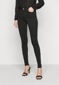 Jeans skinny negros hechos de denim elástico, con una cintura a media altura, diseño de cinco bolsillos y cierre de botón de metal. Llevados con tacones negros.