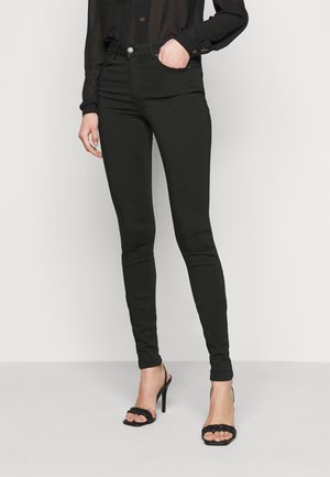 Jeans skinny negros hechos de denim elástico, con una cintura a media altura, diseño de cinco bolsillos y cierre de botón de metal. Llevados con tacones negros.