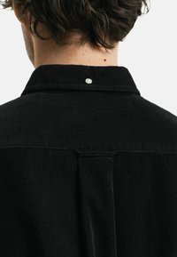 Zwarte corduroy overhemden met een gestructureerde kraag, rugplooidetail en een enkele knoop aan de basis van de kraag. Textuur van de stof zichtbaar.