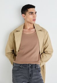 Beige trenchcoat päällä vaaleanruskea ribbimäinen toppi. Tummanharmaat farkut, joissa on hienovaraisia tekstuuridetaljeja. Malli seisoo vaaleaa taustaa vasten.