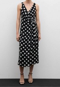 Robe portefeuille à pois noir et blanc avec un décolleté en V, sans manches, taille cintrée et longueur midi, associée à des sandales noires à lanières.