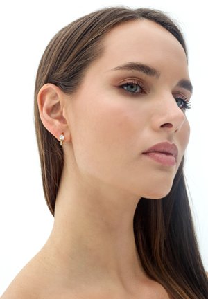 Goldfarbene Ohrringe mit einem klaren Tropfenstein, die ein spiralförmiges Design und strukturierte Akzente aufweisen, getragen von einem Modell mit langen Haaren.