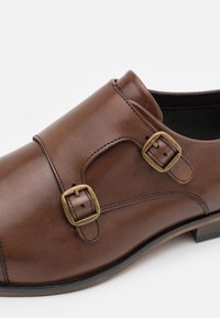 Zapato de cuero marrón con dos hebillas de bronce, textura suave, punta redondeada y tacón bajo, con costuras detalladas a lo largo de las juntas.