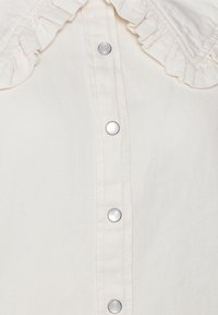 Camicia bianca con colletto volant, bottoni a pressione argentati e un tessuto morbido e texturizzato. Vista ravvicinata che enfatizza i dettagli.