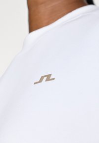 Biela textilná tričko s malým zlatým logom. Okrúhly výstrih a hladká štruktúra, zdôrazňujúce minimalistickú a modernú estetiku.