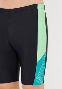 Čierne plávacie šortky s svetlozeleným bočným panelom so zigzag vzorom a bielym logom "Speedo". Hladký, pružný materiál.