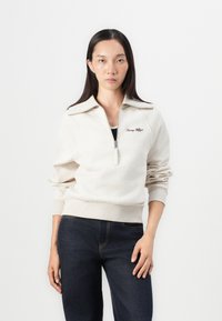 Beige zip-up sweatshirt with high collar, ribbed cuffs, and hem. Features embroidered "Tommy Hilfiger" logo on the chest. Paired with dark jeans.