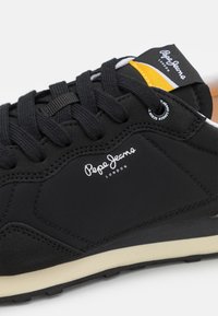 Baskets noires Pepe Jeans avec lacets noirs, logo blanc sur le côté, détail jaune et blanc sur la languette et semelle beige.