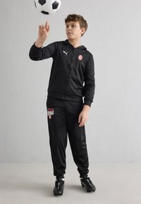 Rapaz vestido com fato de treino preto do AC Milan a fazer rodar uma bola de futebol no dedo, contra um fundo simples de parede cinzenta clara.