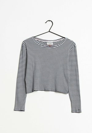 Sort og hvid langærmet cropped skjorte med vandrette striber hængende på en sort bøjle mod en hvid baggrund.