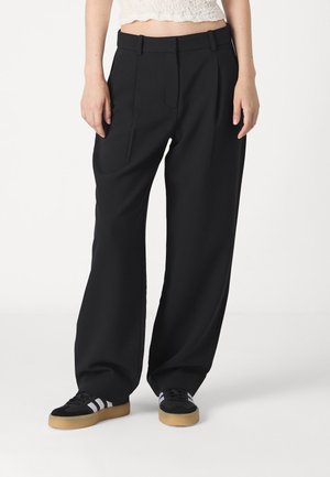 Black wide-leg trousers with a smooth texture, featuring front pleats and side pockets, paired with black sneakers and gum sole accents.