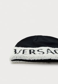 Pletena crna kapa s debelim bijelim čupavim rubom na kojem je urezani crni tekst "VERSACE," prikazujući teksturirani dizajn.