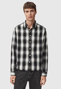 Koszula w czarno-białą kratę z miękkiego materiału, z klasycznym kołnierzykiem, długimi rękawami i zaokrąglonym dołem, z białymi guzikami.