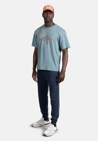 Lyseblå bomulds T-shirt med bjergdesign og orange solgrafik, parret med marineblå sweatpants og grå sportssko. Kasketten er koralrød og blå.