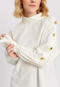 Naine, kes kannab valget pika varrukaga särki, mille õlgadel ja varrukatel on kuldsed nööbid, kohandab mansetti mõlema käega.