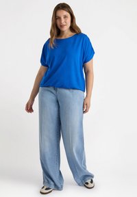 Blaues Kurzarmoberteil mit lockerer Passform, kombiniert mit hellblauen weiten Jeans und schwarz-weißen Sneakern. Glatter Stoff, minimalistisches Design.