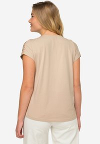 Camiseta de manga corta beige con detalle de cordones en el hombro, textura suave y un corte holgado, combinada con pantalones de color claro.