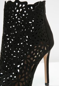 Bottine noire à la cheville avec un motif floral découpé au laser, en matériau suédé, talon stiletto haut et design à bord festonné, mettant en valeur un travail détaillé et raffiné.