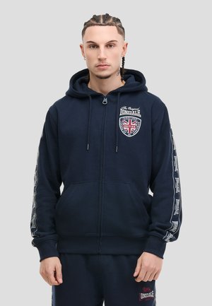 Ung man med flätat hår som bär en marinblå Lonsdale-hoodie med dragkedja och matchande mjukisbyxor mot en enkel ljus bakgrund.