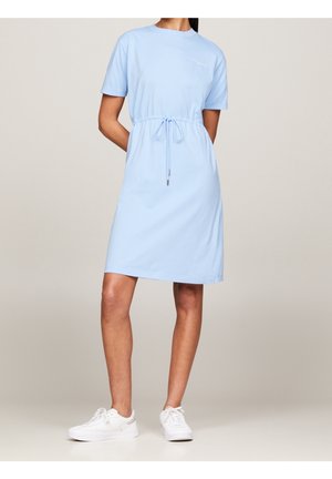 Femme portant une robe bleu clair courte aux manches courtes, une ceinture à la taille et des baskets blanches, debout devant un fond uni.