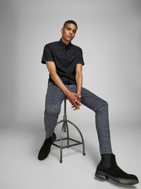 Chemise noire à manches courtes avec boutons, pantalon à carreaux texturé gris, et bottines en daim noires. Assis sur un tabouret en métal devant un fond gris.