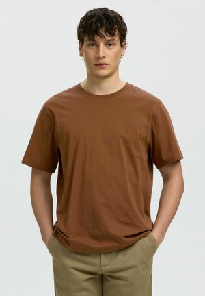 Giovane uomo con capelli scuri e ricci che indossa una maglietta marrone a maniche corte e pantaloni beige, in piedi con le mani nelle tasche davanti a uno sfondo semplice.
