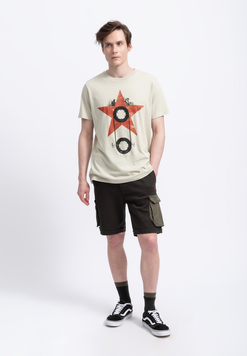 Beige t-shirt met een rode ster graphic en zwarte cirkelvormige accenten, gecombineerd met donkere shorts met cargozakken en zwarte sneakers.