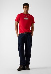 Czerwona bawełniana koszulka z logo "GUESS JEANS", w połączeniu z ciemnoniebieskimi spodniami typu cargo z bocznymi kieszeniami oraz brązowymi butami trekkingowymi.