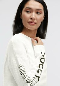 Weißer Strickpullover mit Rippenstruktur, mit schwarzem "soccer"-Schriftzug am linken Ärmel und rundem Halsausschnitt.