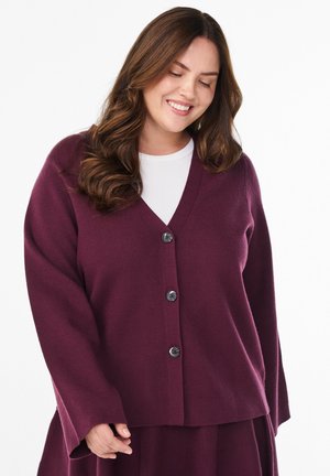 Kobieta z długimi brązowymi włosami, ubrana w zapinaną na guziki kardigan w kolorze śliwkowym na białej bluzce, uśmiechająca się i patrząca w dół na jednolitym tle.