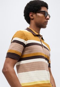 Άνδρας με αφρο κούρεμα που φοράει μαύρα γυαλιά ηλίου και κοντομάνικο πλεκτό πόλο με οριζόντιες ρίγες σε καφέ, κίτρινο, μπεζ και λευκό χρώμα.